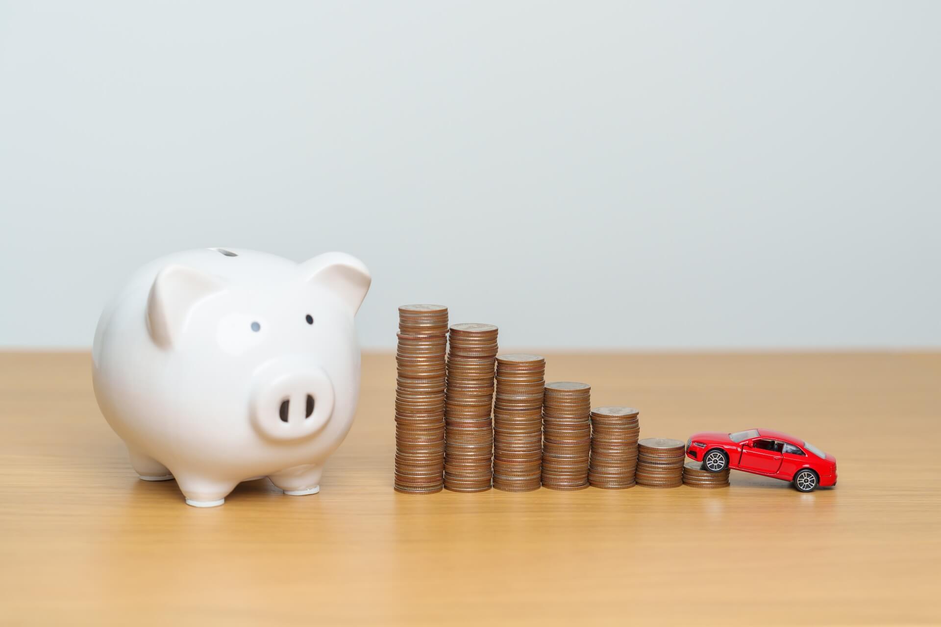 White piggy bank to the left of piles of coins with a small red toy car on top o the smallest stack of coins | Featured image for the What Is a Good Car Finance Rate blog by FinanceBeagle.